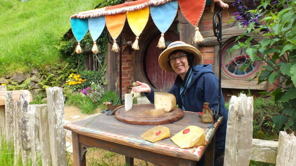 An image of Jean pretending to offer cheese for sale at a Hobbit hole in Hobbiton in Matamata, New Zealand. Photography by Frame To Frame - Bob and Jean